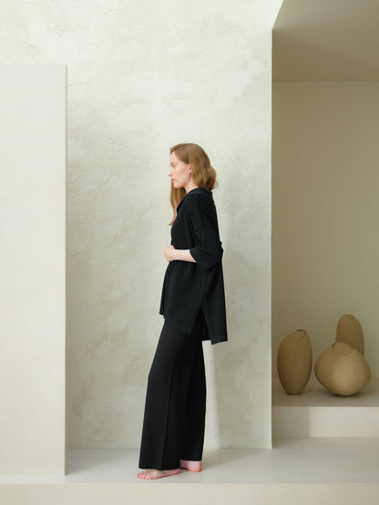 Profile view of a model wearing a black Extra Fine Merino Wool Maxi Polo with wide-leg trousers against a pale textured backdrop.
