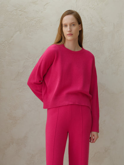 Bright pink cashmere mix crewneck sweater worn by a model against a textured beige wall.