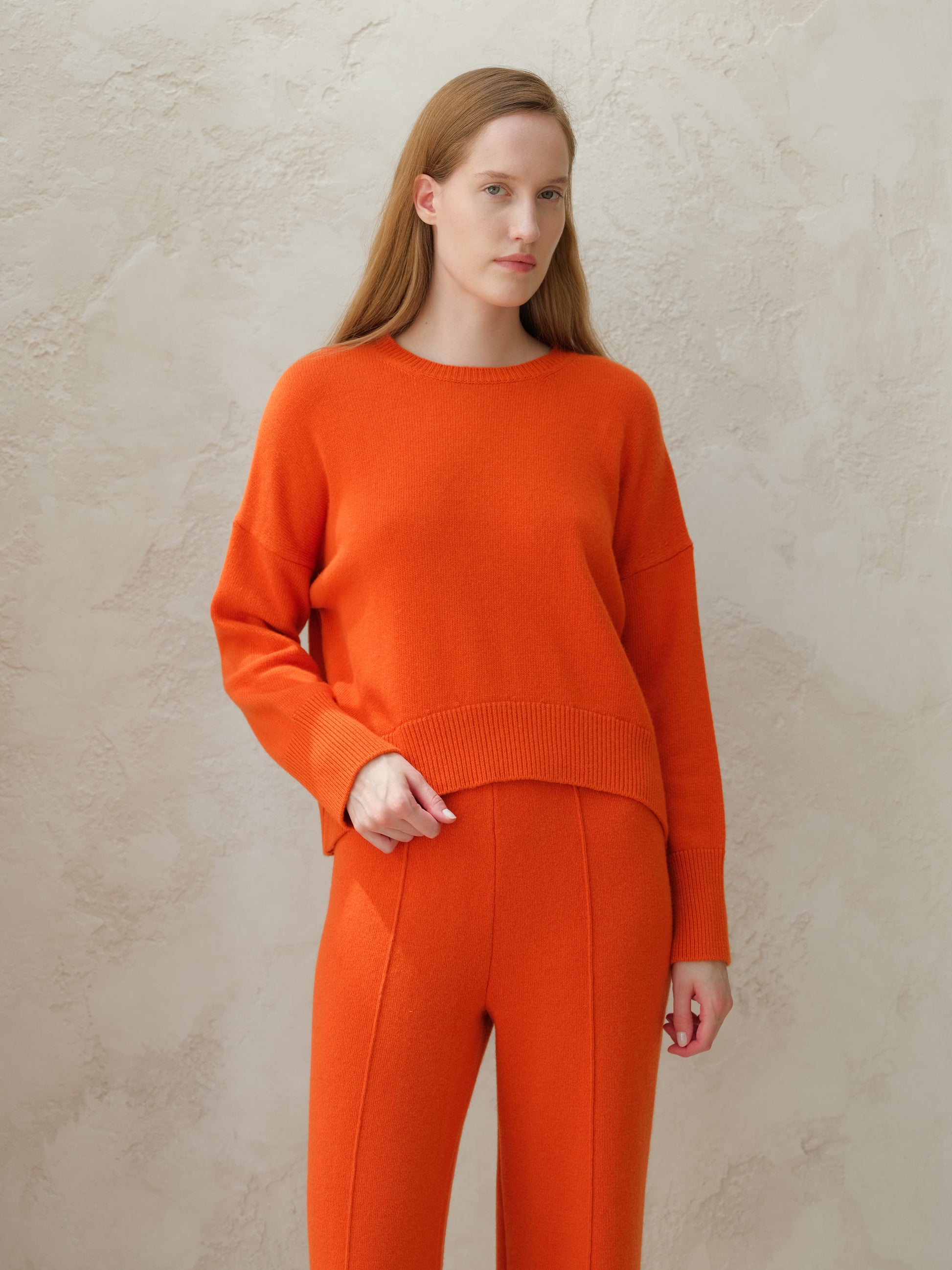 Model wearing orange cashmere mix crewneck sweater with matching orange knit pants against a textured wall.