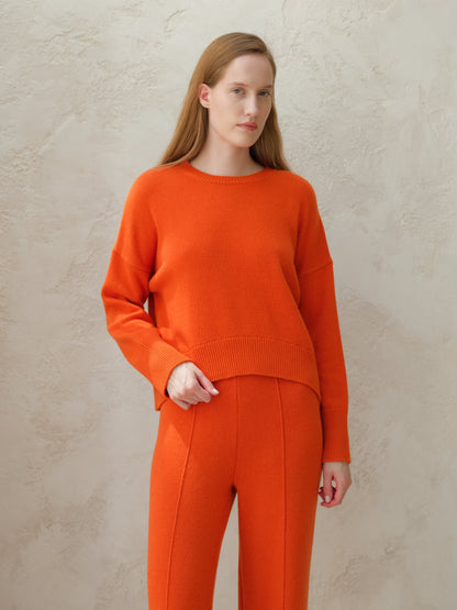 Model wearing orange cashmere mix crewneck sweater with matching orange knit pants against a textured wall.