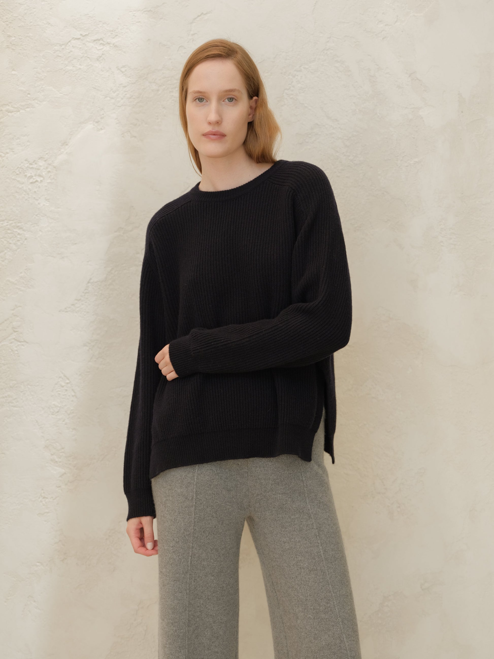 Model wearing black cashmere mix crewneck ribbed sweater with side slits and gray pants against a textured wall