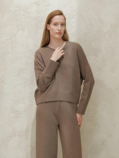 Model wearing taupe cashmere mix crewneck sweater with matching trousers against a pale textured wall
