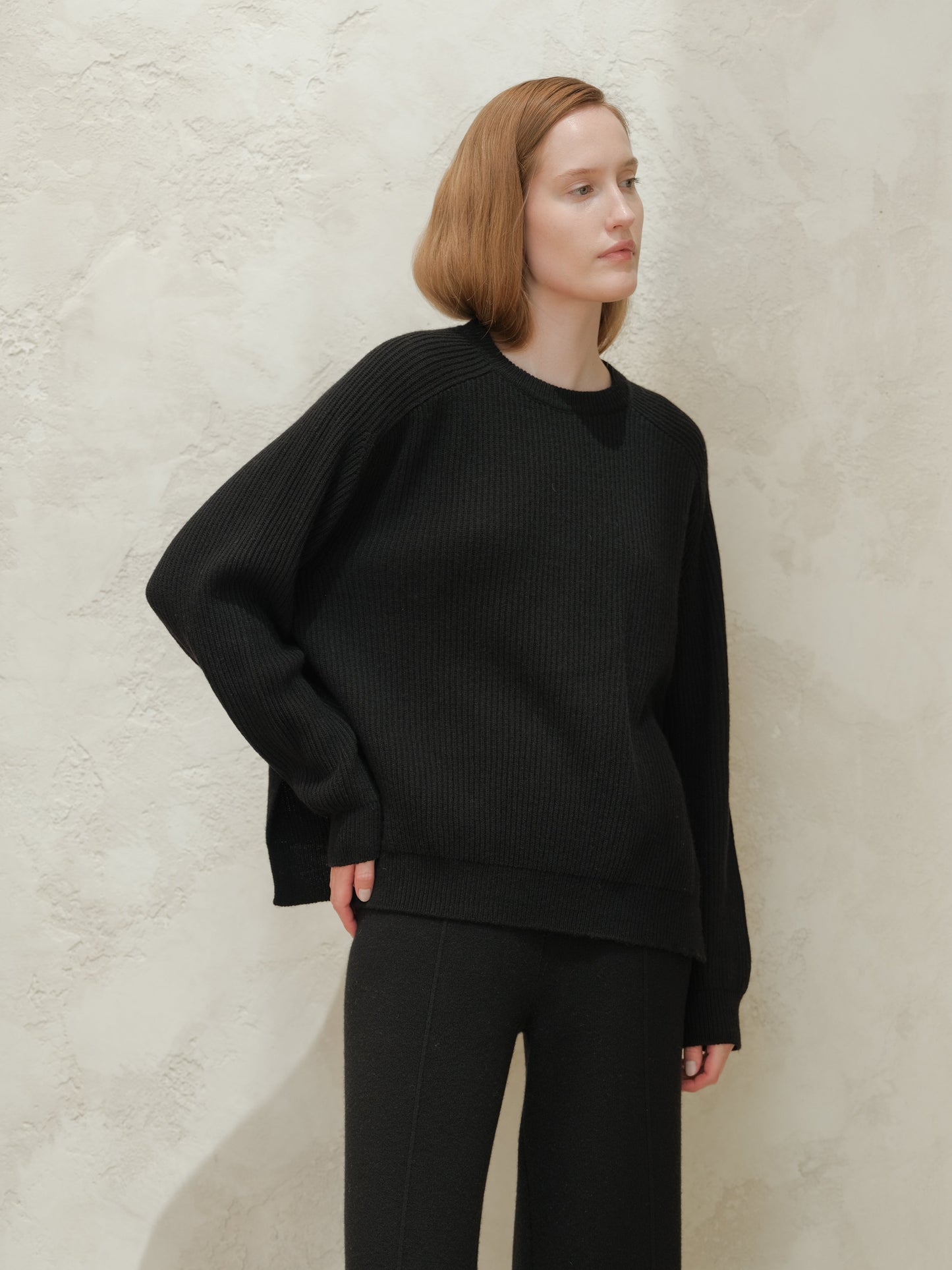 Model wearing black cashmere mix crewneck ribbed sweater with side slits against a textured light wall