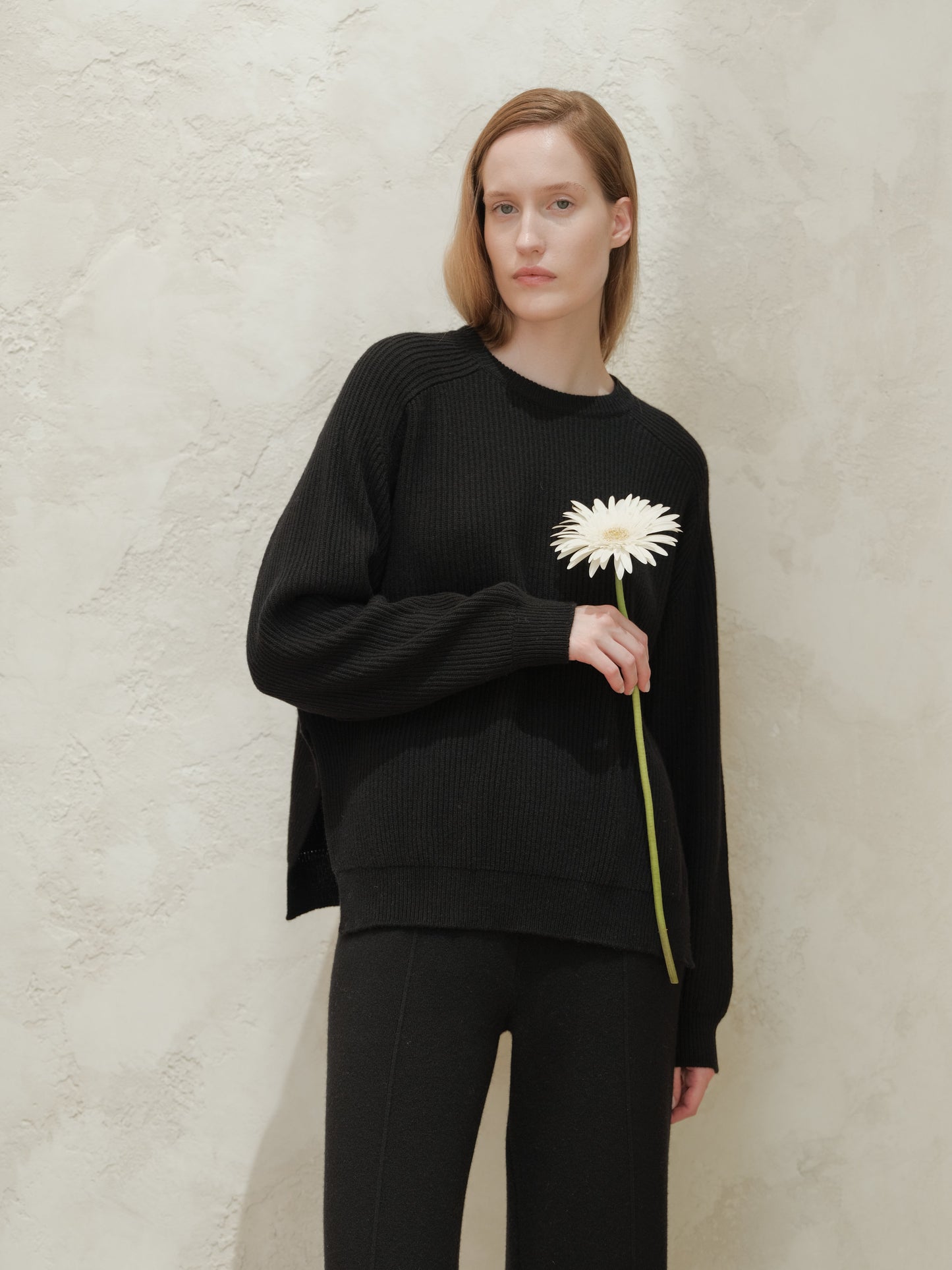 Model wearing a black ribbed cashmere mix crewneck sweater with side slits holding a white flower against a textured beige wall.