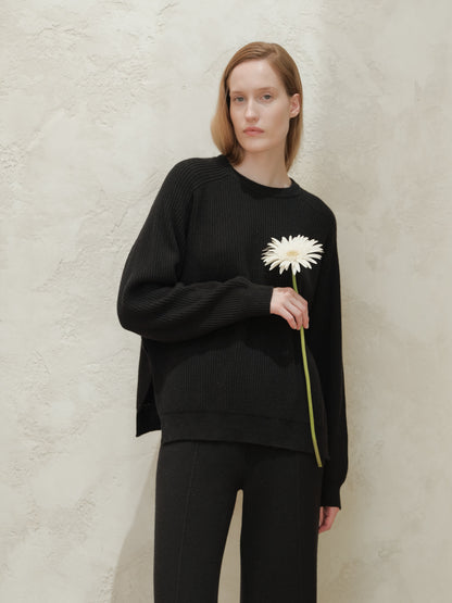 Model wearing a black ribbed cashmere mix crewneck sweater with side slits holding a white flower against a textured beige wall.