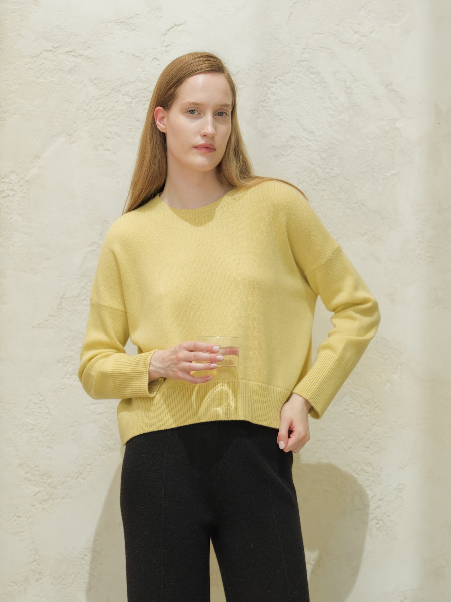 Model in pale yellow cashmere mix crewneck sweater with black pants against a textured wall.