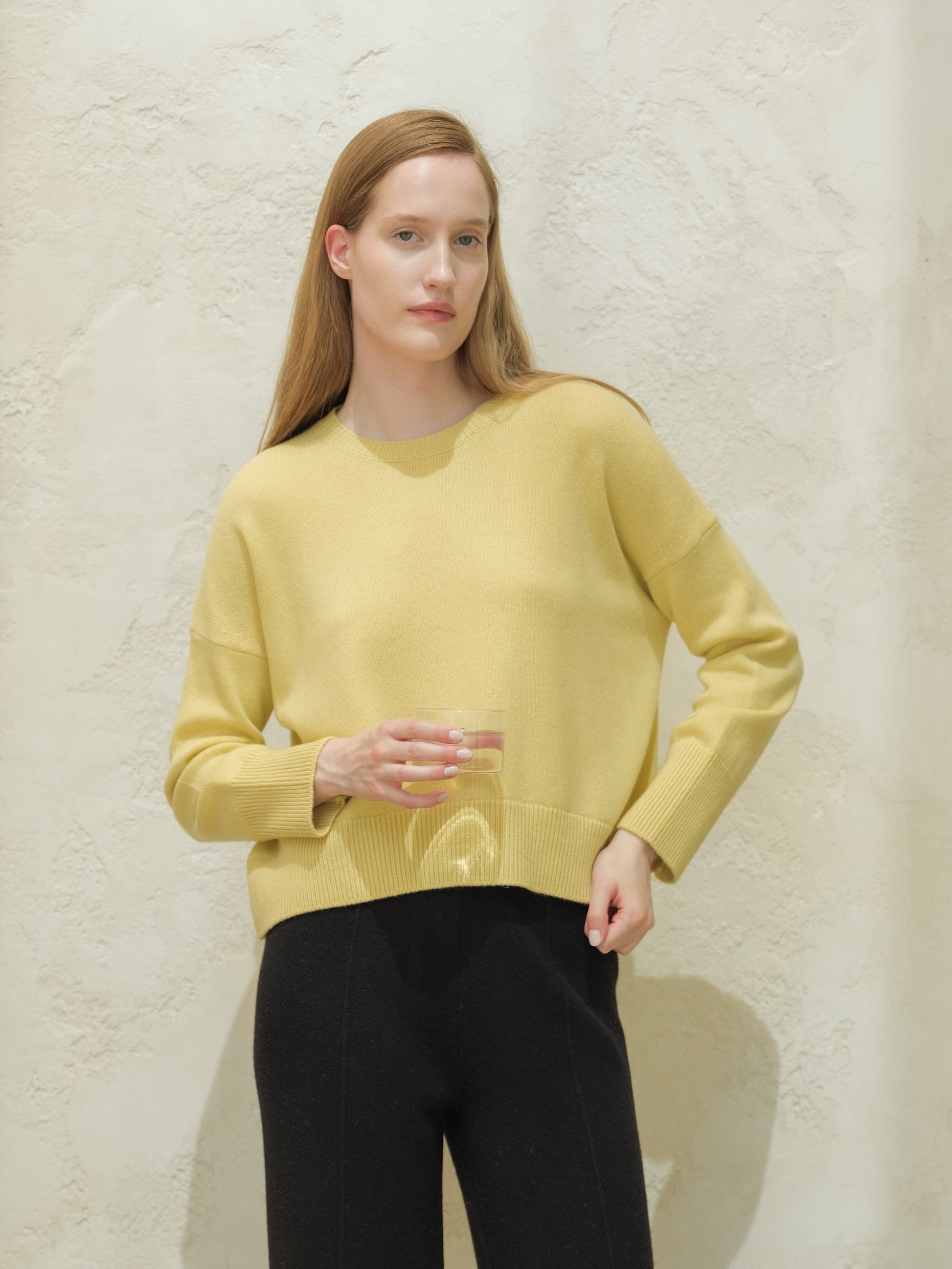 Model in pale yellow cashmere mix crewneck sweater with black pants against a textured wall.