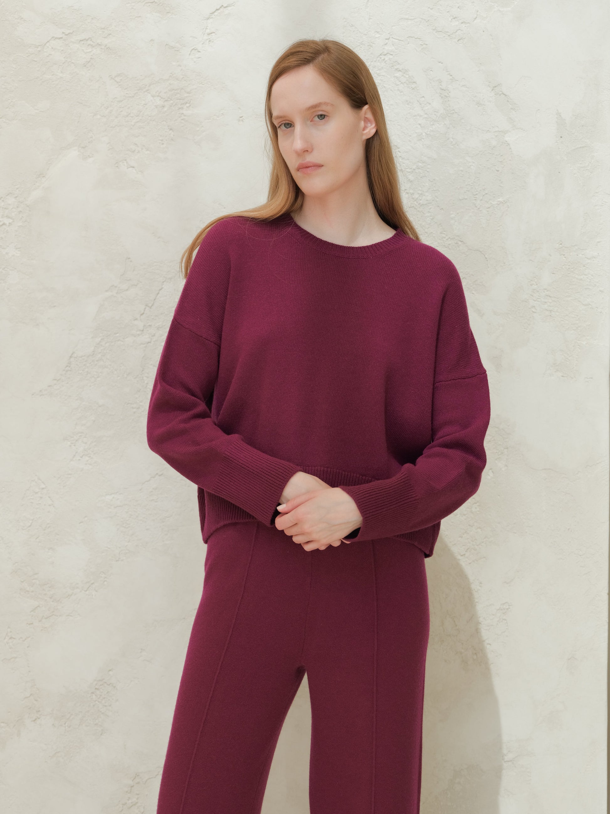 Model wearing a deep burgundy cashmere mix crewneck sweater with matching knit pants against a textured light wall