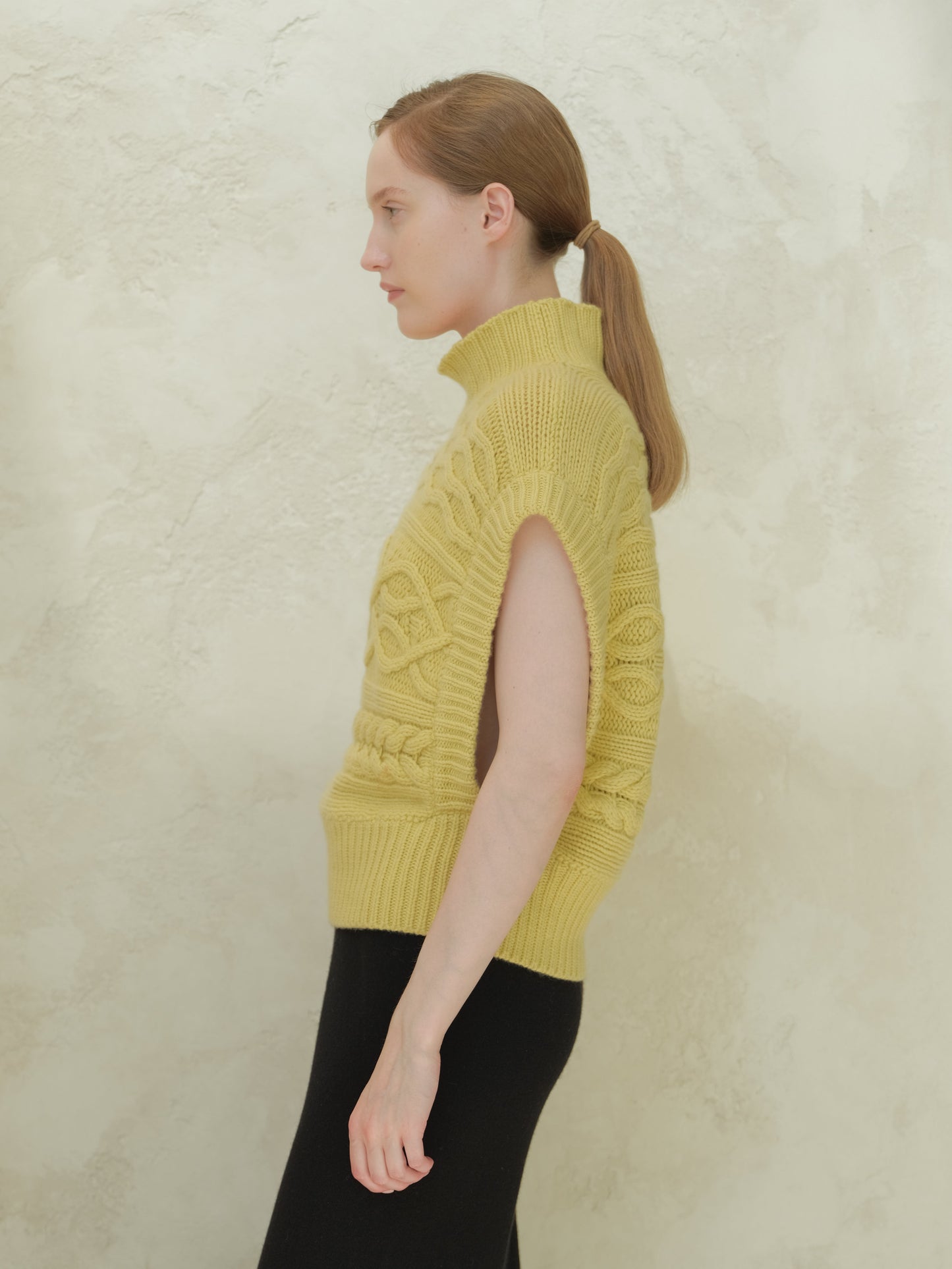 Side view of a model wearing a pale yellow high neck cable knit maxi tunic with sleeveless silhouette.