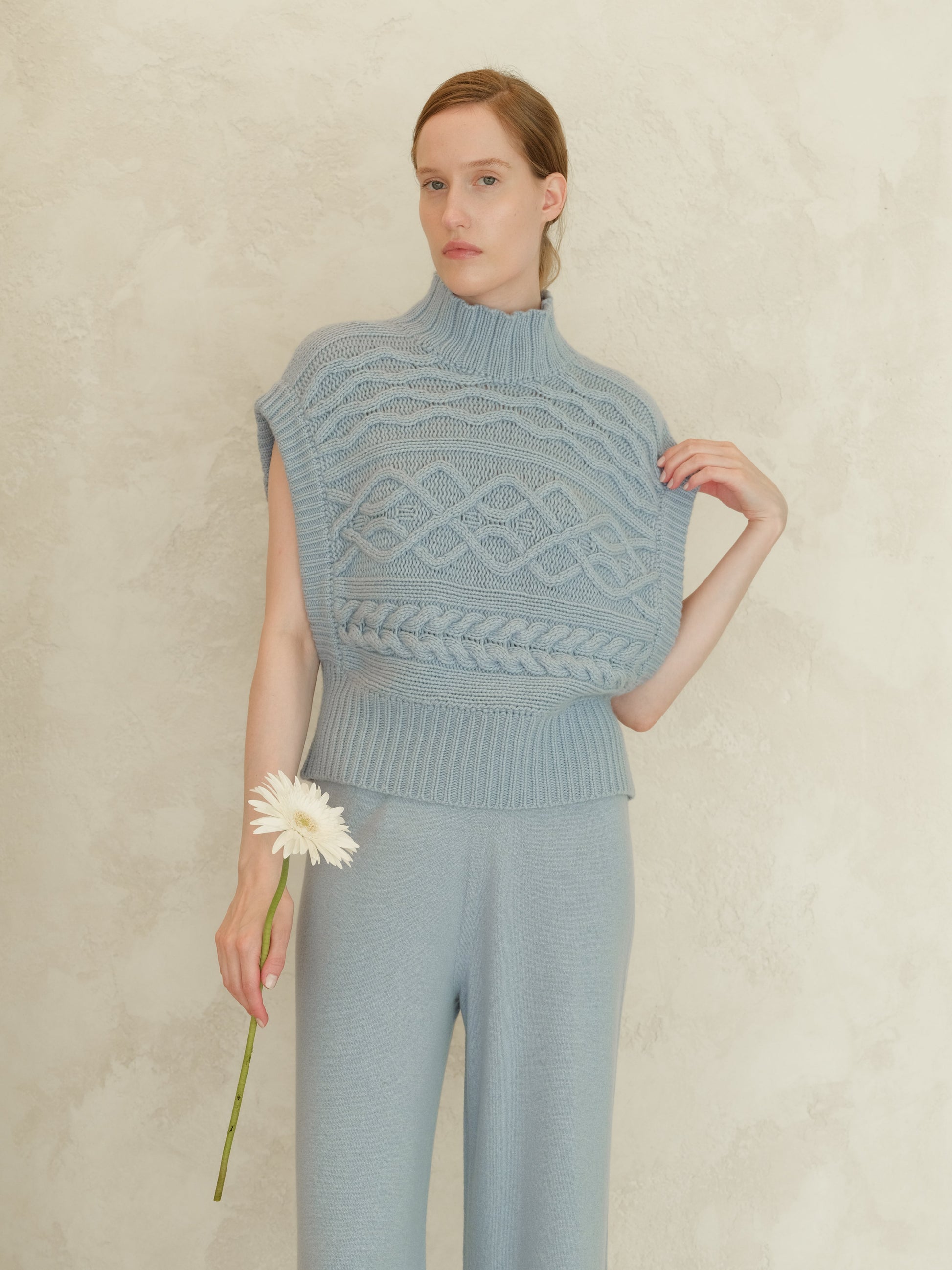 Model wearing blue cashmere mix maxi cable tunic with a high ribbed neck and sleeveless silhouette against a textured wall.