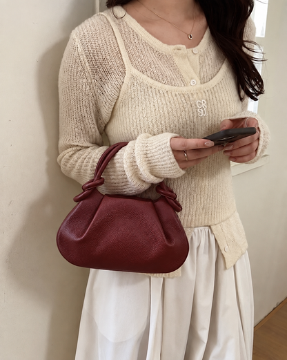 A person wearing a cream ribbed knit cardigan over a white camisole and white skirt, holding a dark phone and carrying a small pebbled leather shoulder bag in a muted burgundy red. The cardigan has a small silver GG-style monogram on the chest.