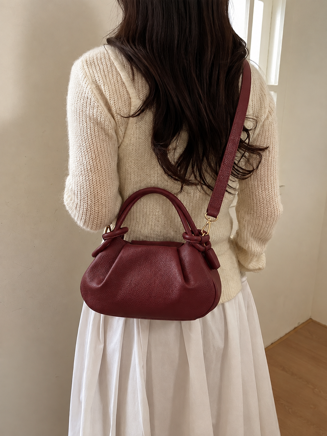 A woman wearing a cream knit sweater and white skirt carries a small burgundy leather handbag with a top handle and shoulder strap, shown from behind in an indoor setting.