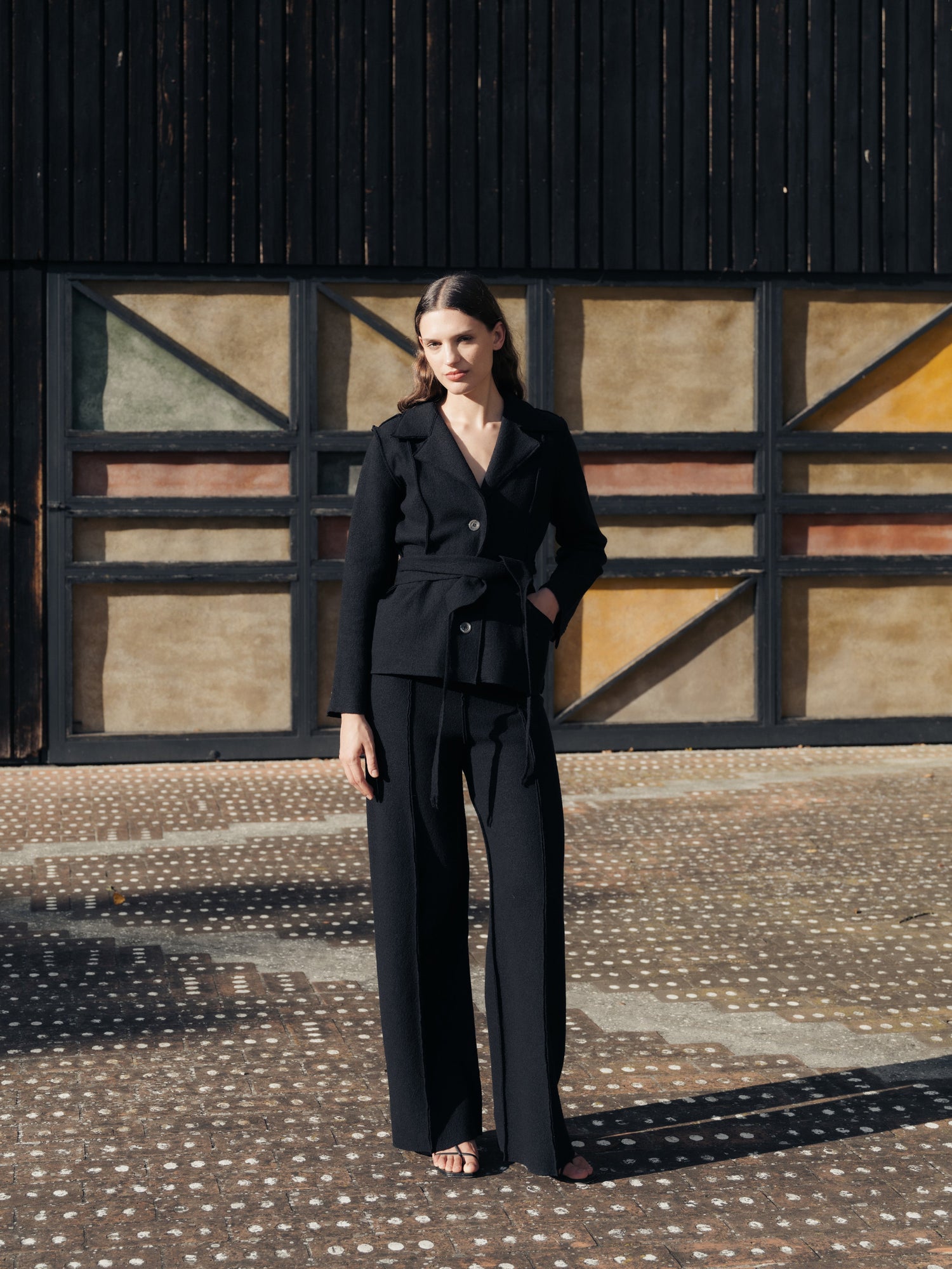 Woman in a black boiled wool suit standing in front of a colorful geometric-patterned wall.