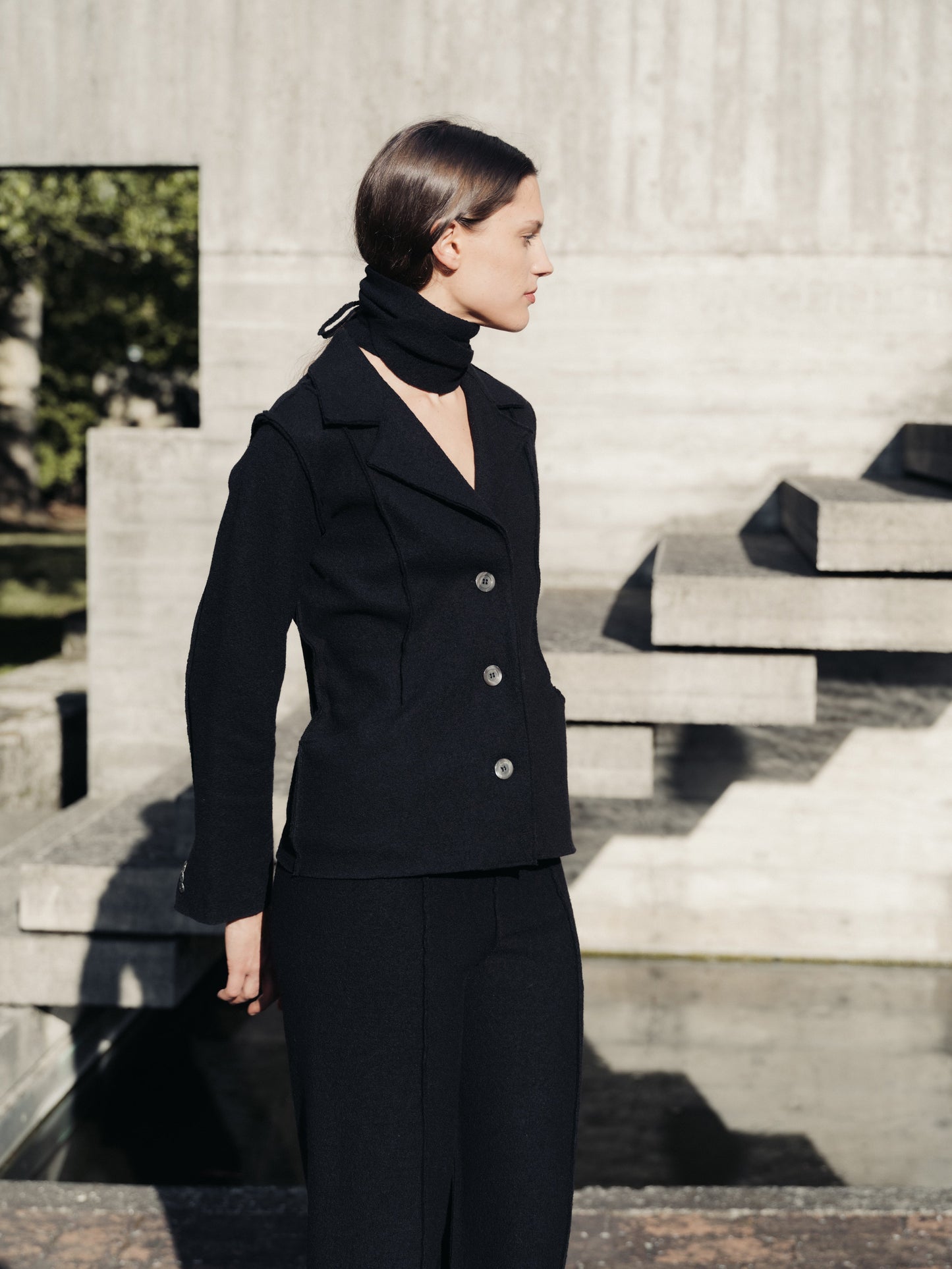 Woman in a black boiled wool suit standing outdoors on stone steps.