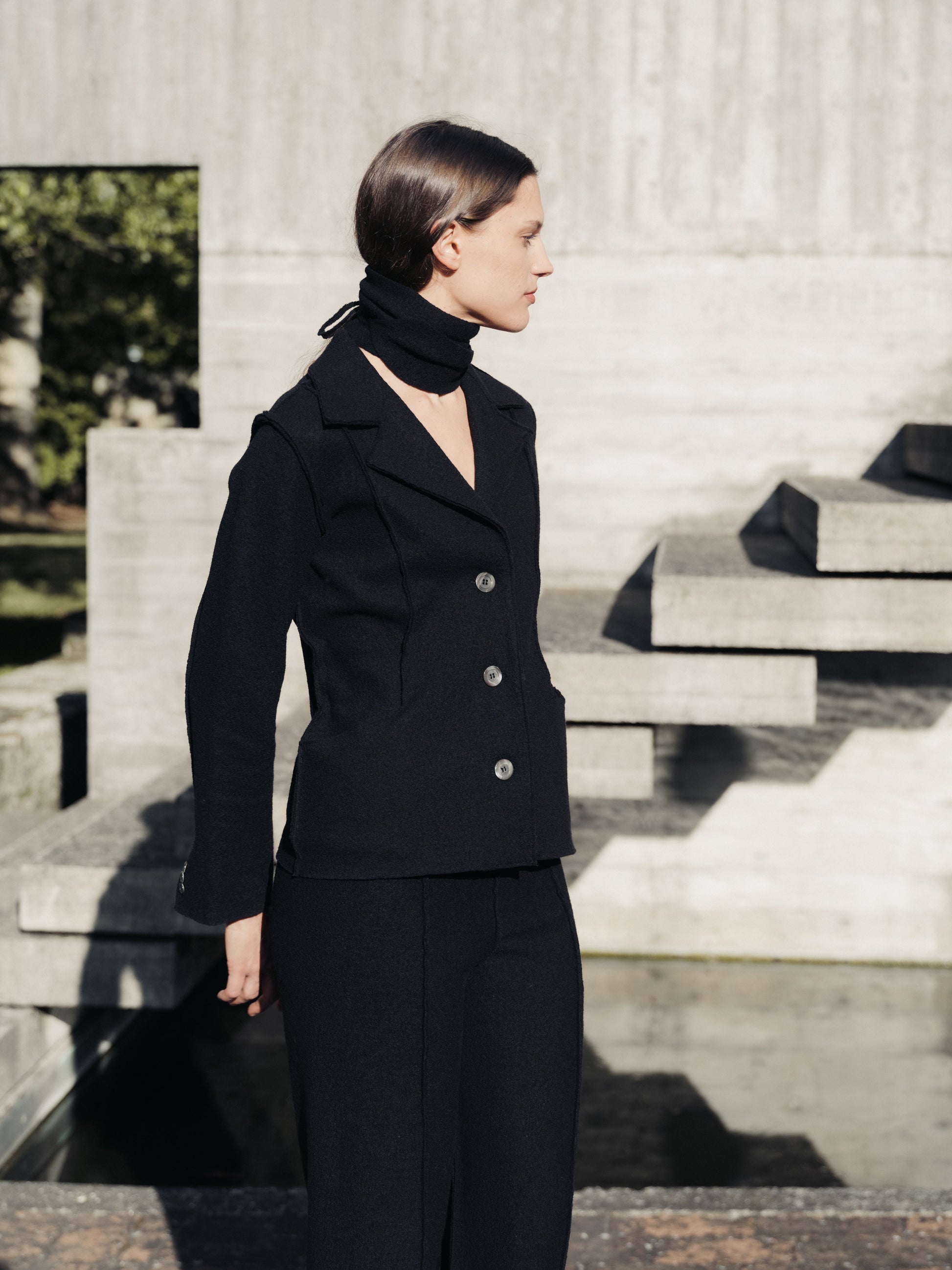 Woman in a black boiled wool suit standing outdoors on stone steps.