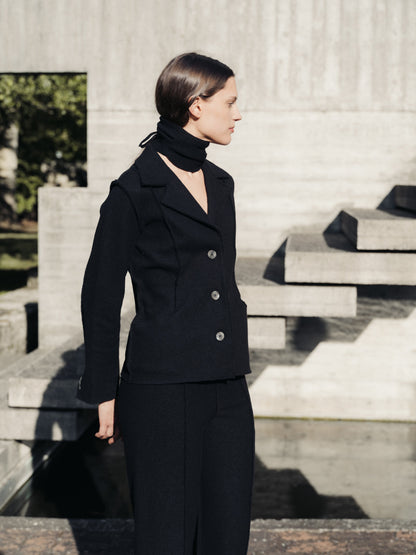 Woman in a black boiled wool suit standing outdoors on stone steps.