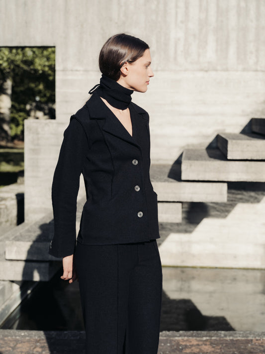 Woman in a black boiled wool suit standing outdoors on stone steps.