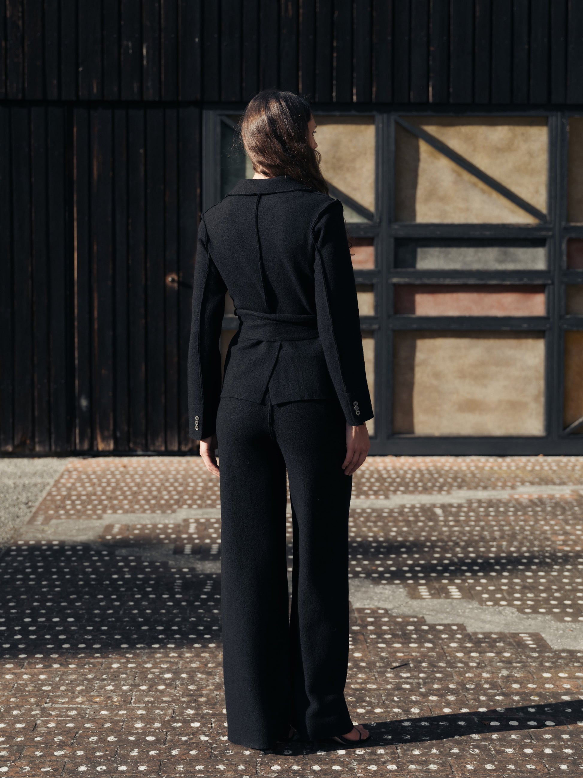 Back view of woman wearing a black boiled wool suit standing in front of a rustic wooden door.