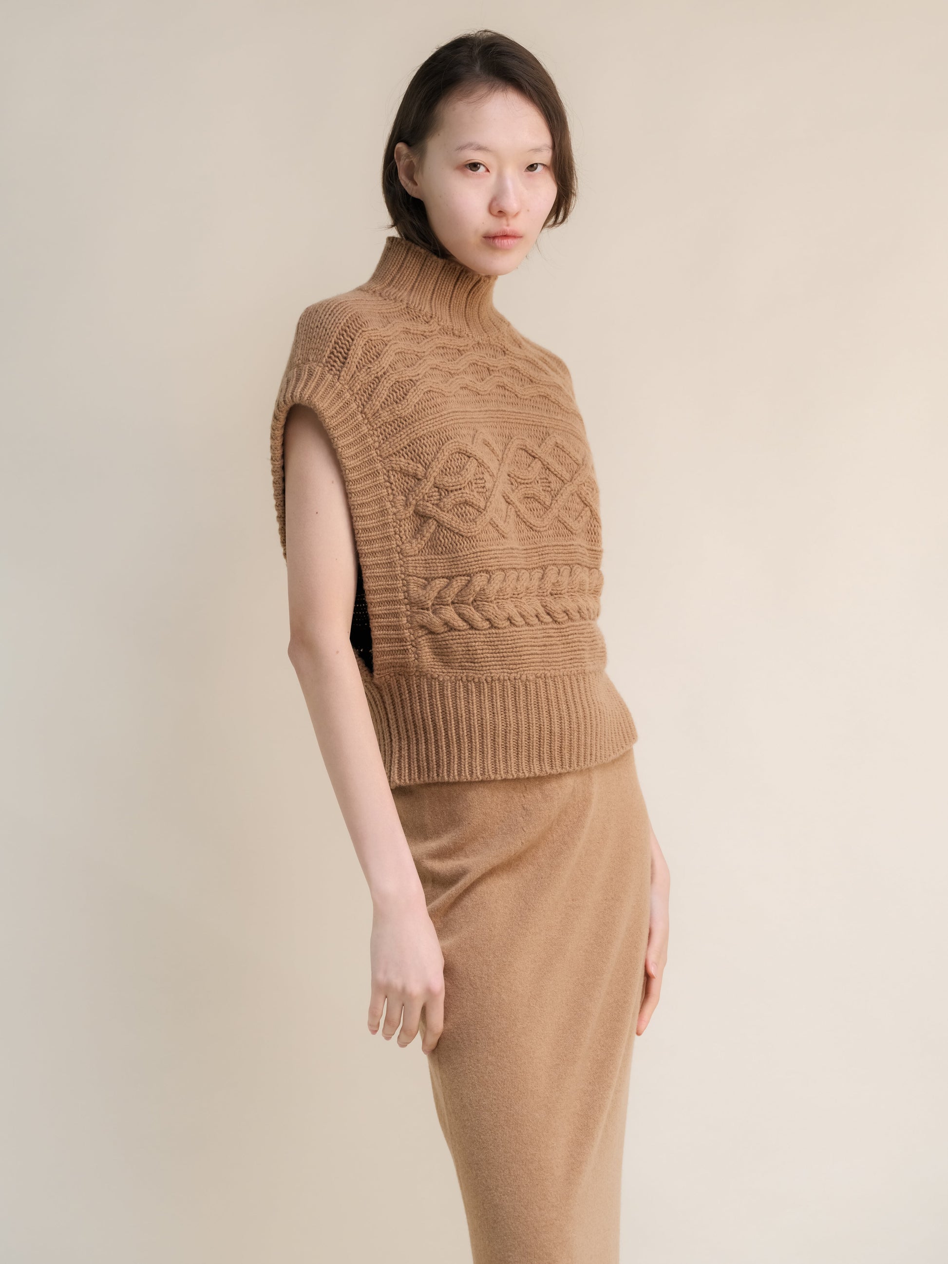 Beige knit sleeveless turtleneck tunic with bold cable patterns worn with a matching camel skirt.