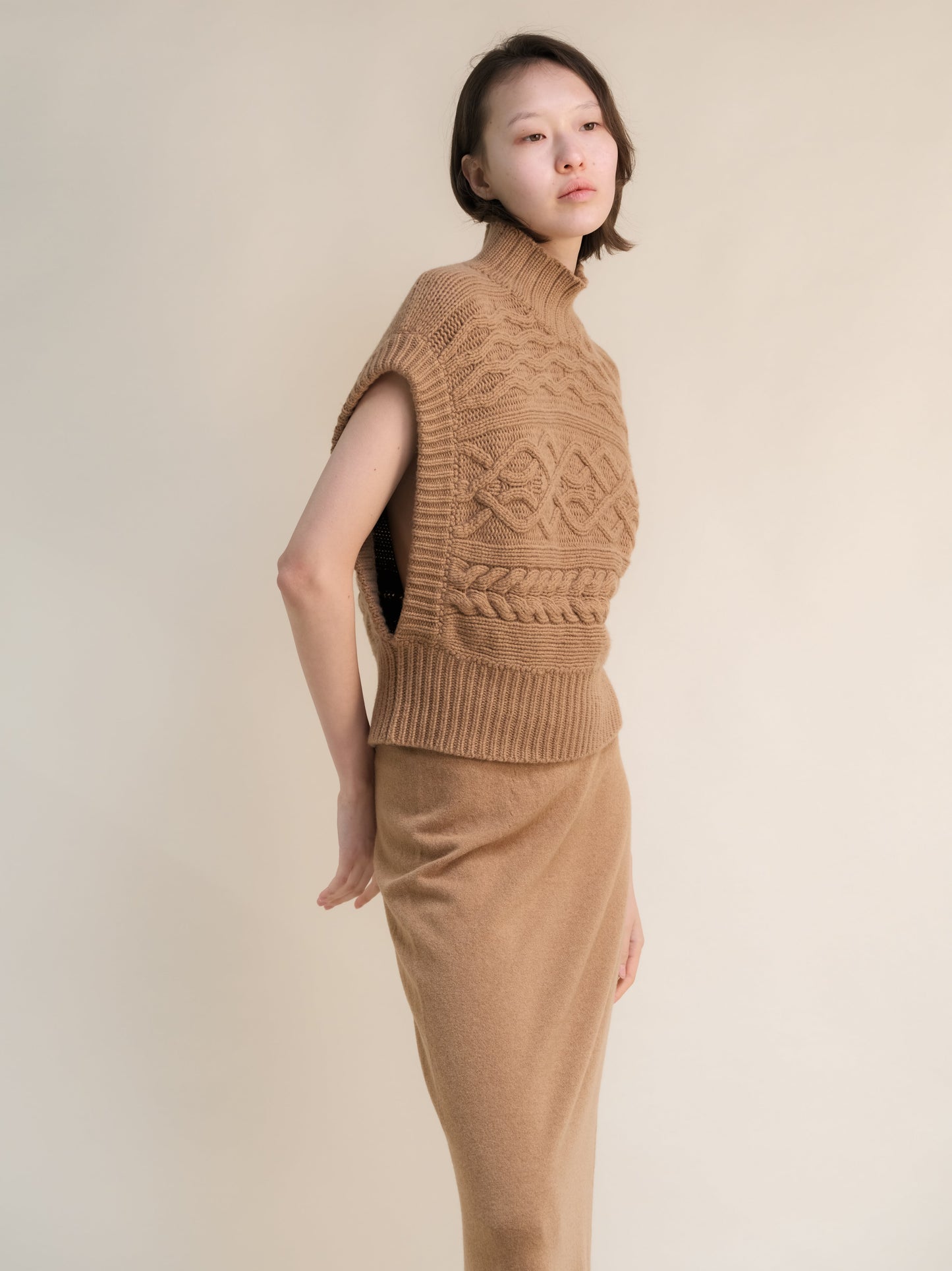 Profile view of a model wearing a tan cashmere mix maxi cable tunic with a high ribbed neck and bold knit patterns.
