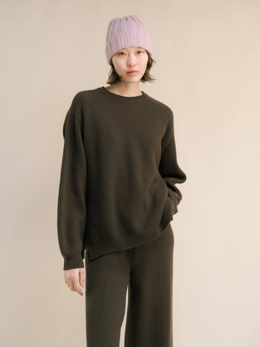 Person wearing a dark green sweater and pants with a light purple knit beanie on a beige background