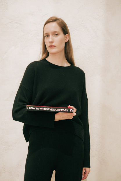 Model wearing a black cashmere mix crewneck sweater holding a red book at the hip