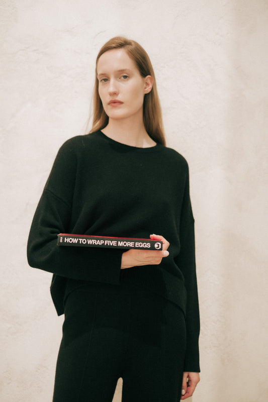 Model wearing a black cashmere mix crewneck sweater holding a red book at the hip