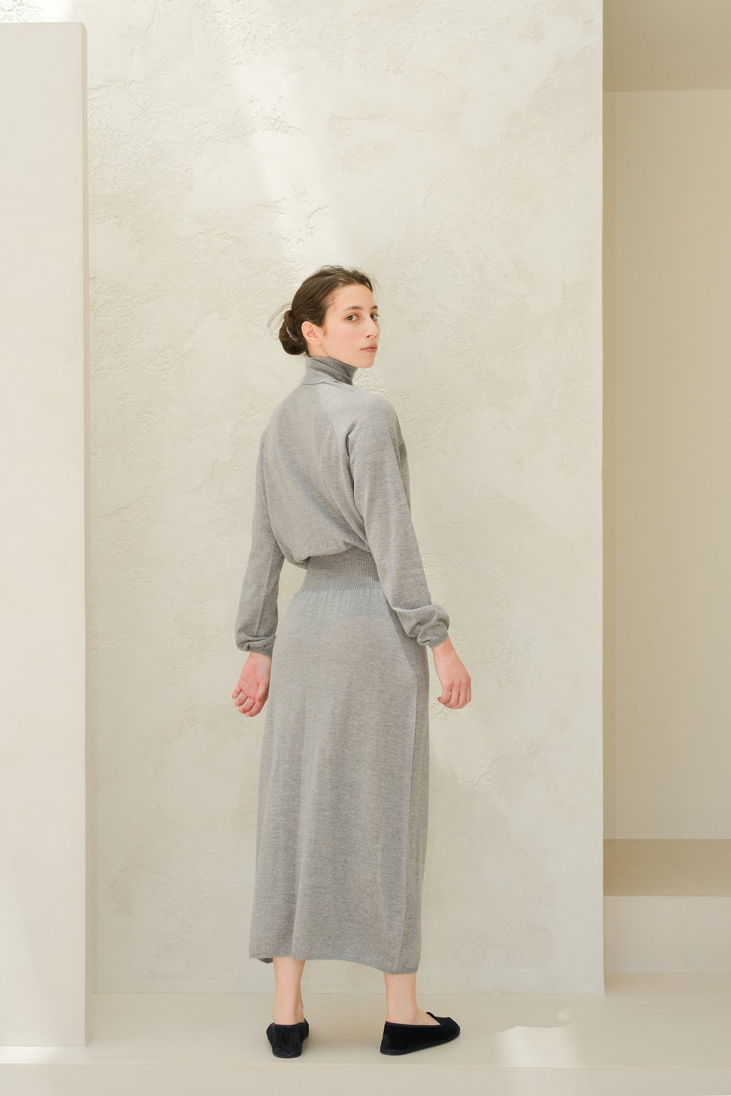 Model wearing a light grey 3D knit merino wool dress from behind with a cinched waist and long sleeves.