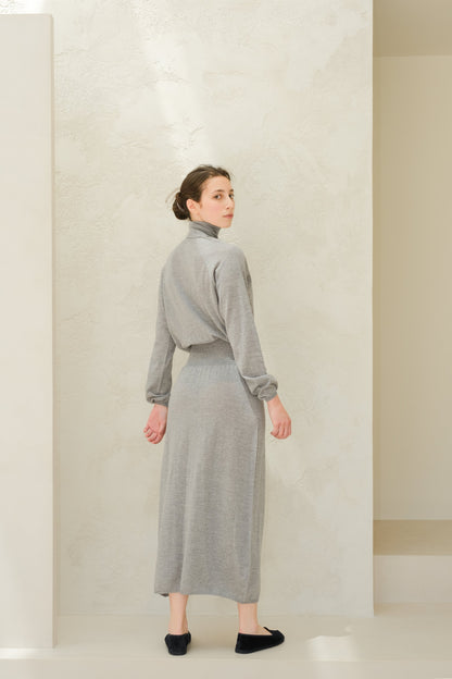 Model wearing a light grey 3D knit merino wool dress from behind with a cinched waist and long sleeves.