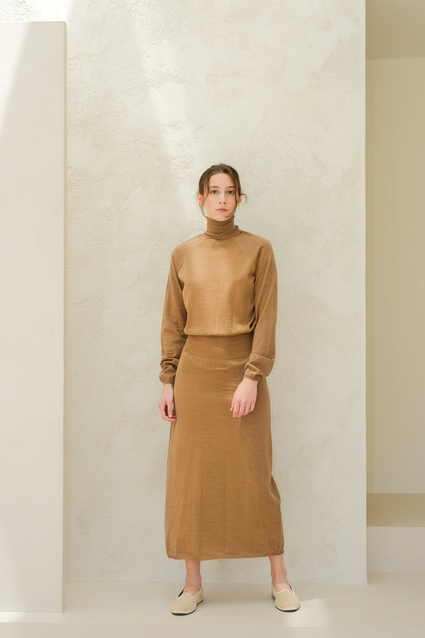 Full-body view of a model in camel brown 3D knit merino dress set with a turtleneck top and ribbed-waist skirt, beige slip-on shoes
