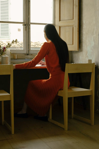 Woman in an orange pleated knit skirt and orange sweater sitting at a table by a window in a dimly lit room.