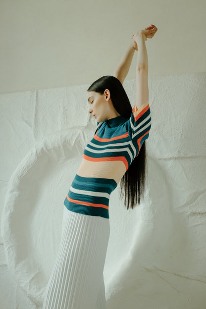 Profile of a model in a multicolor striped knit top and a white pleated long skirt, arms raised overhead.