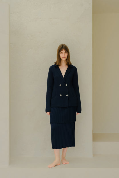 Navy knitted double breasted blazer with a layered pleated long skirt on a barefoot model against a beige textured wall