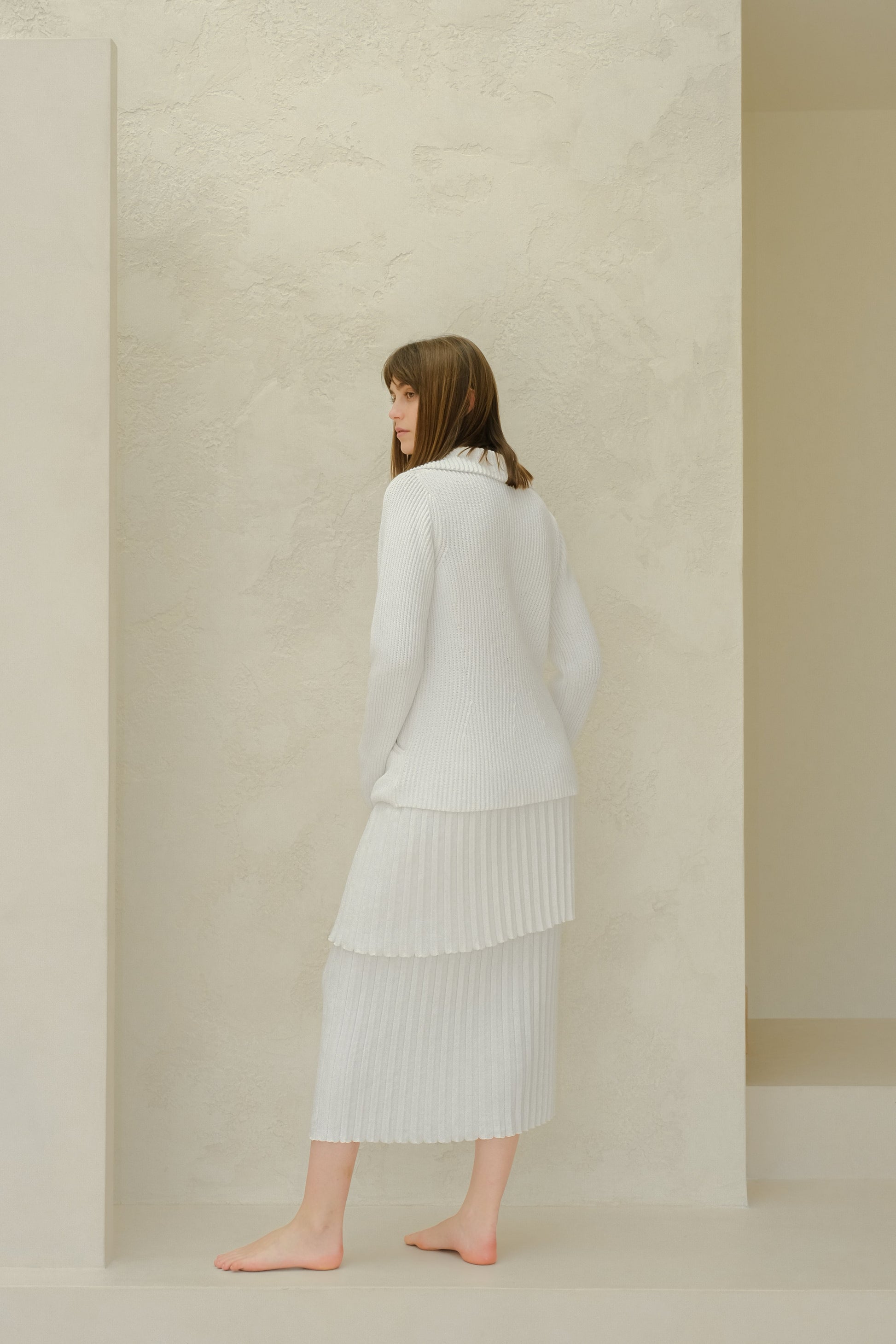 Side view of a model wearing a white fine cotton plissé knit skirt with a matching sweater against a textured beige wall.