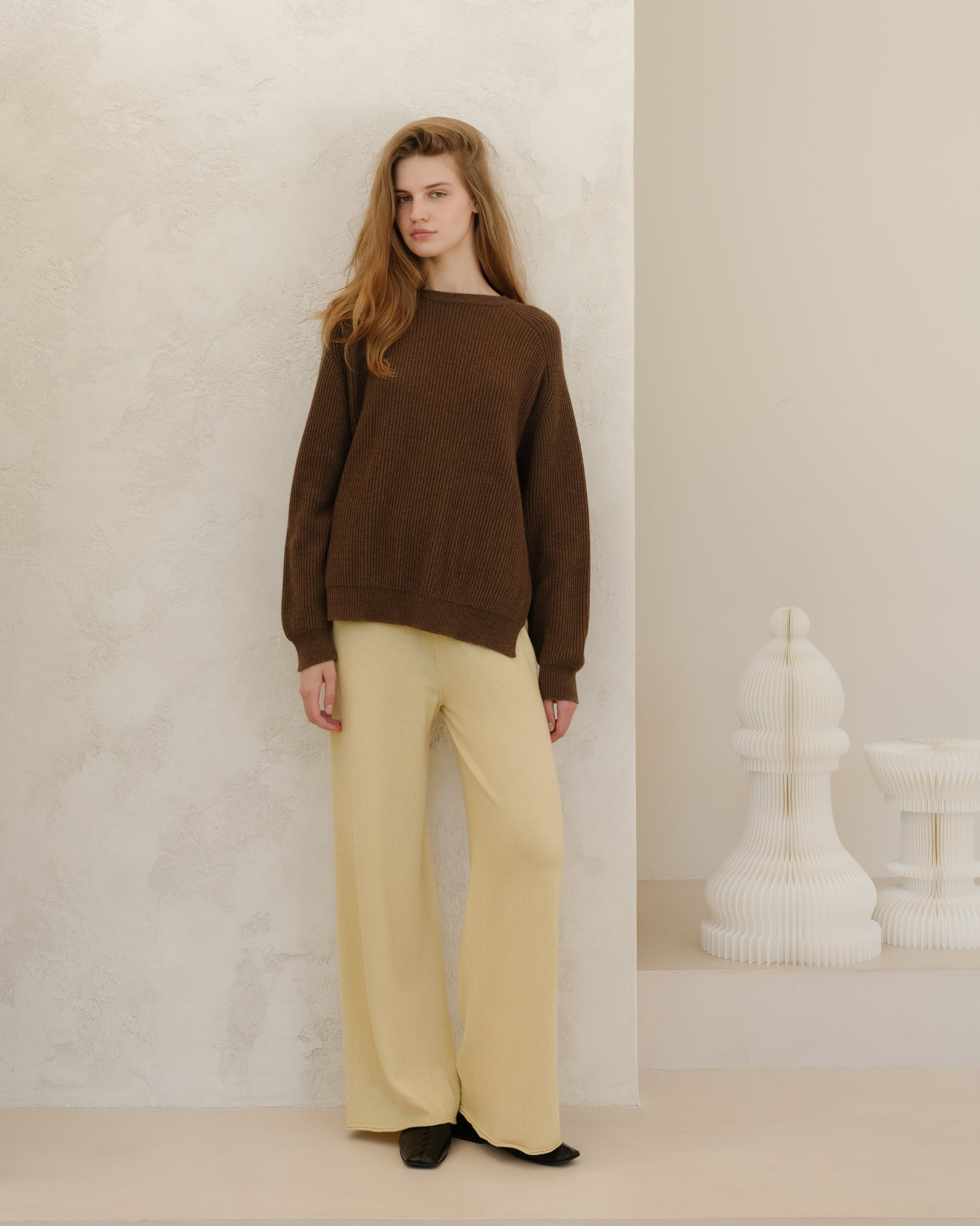 Model wearing brown ribbed cashmere mix crewneck sweater with lateral slits, paired with pale beige wide-leg pants