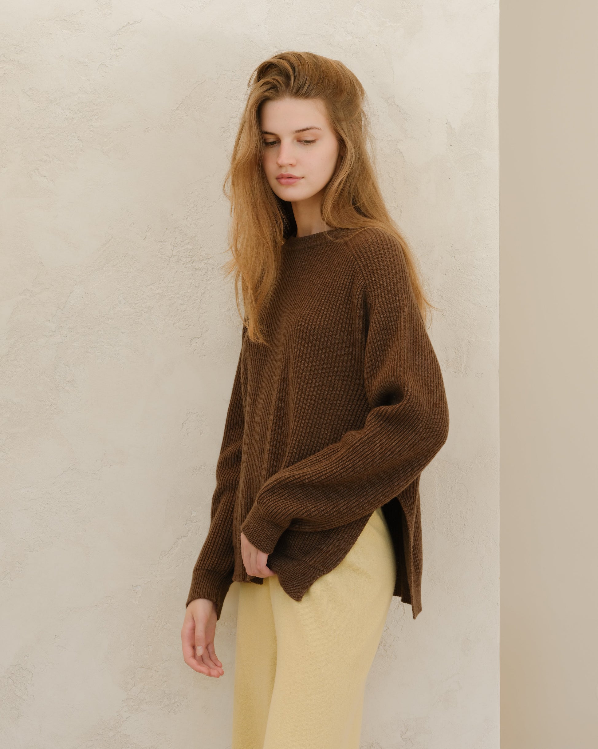 Model in a brown ribbed knit crewneck sweater with side slits, paired with beige pants, standing against a textured wall.