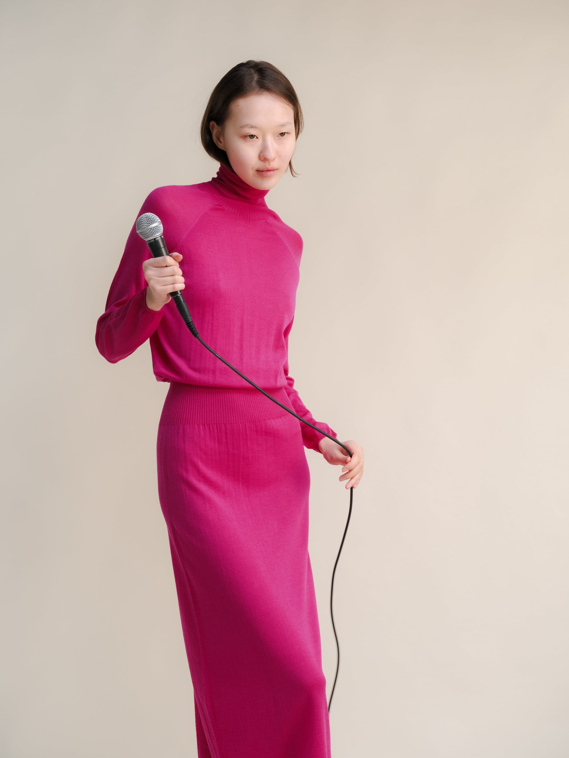 Model in magenta 3D knit merino wool dress holding a microphone against a pale studio backdrop