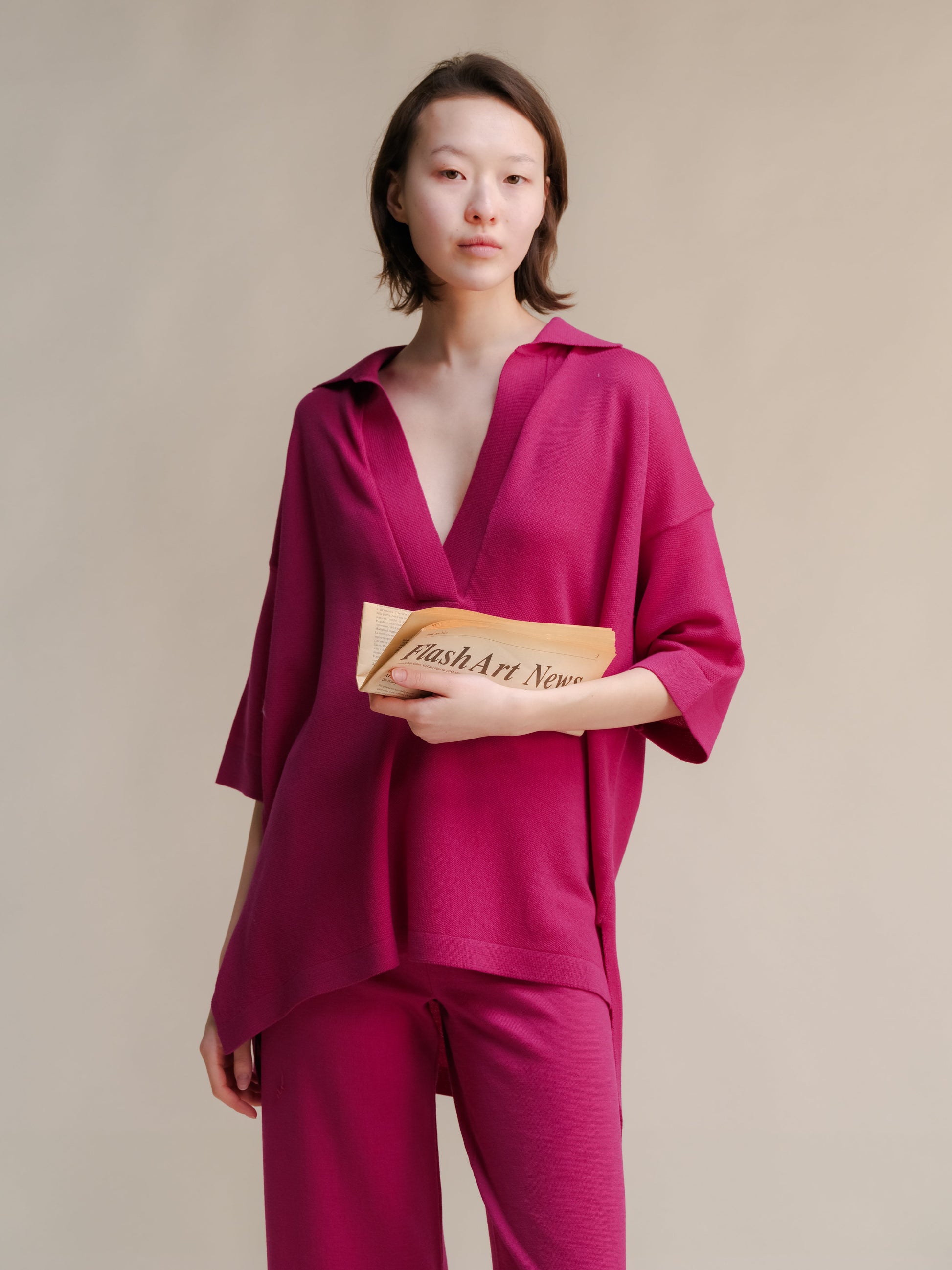 Bright pink merino wool maxi polo worn by a model with a deep V neckline, holding a folded newspaper