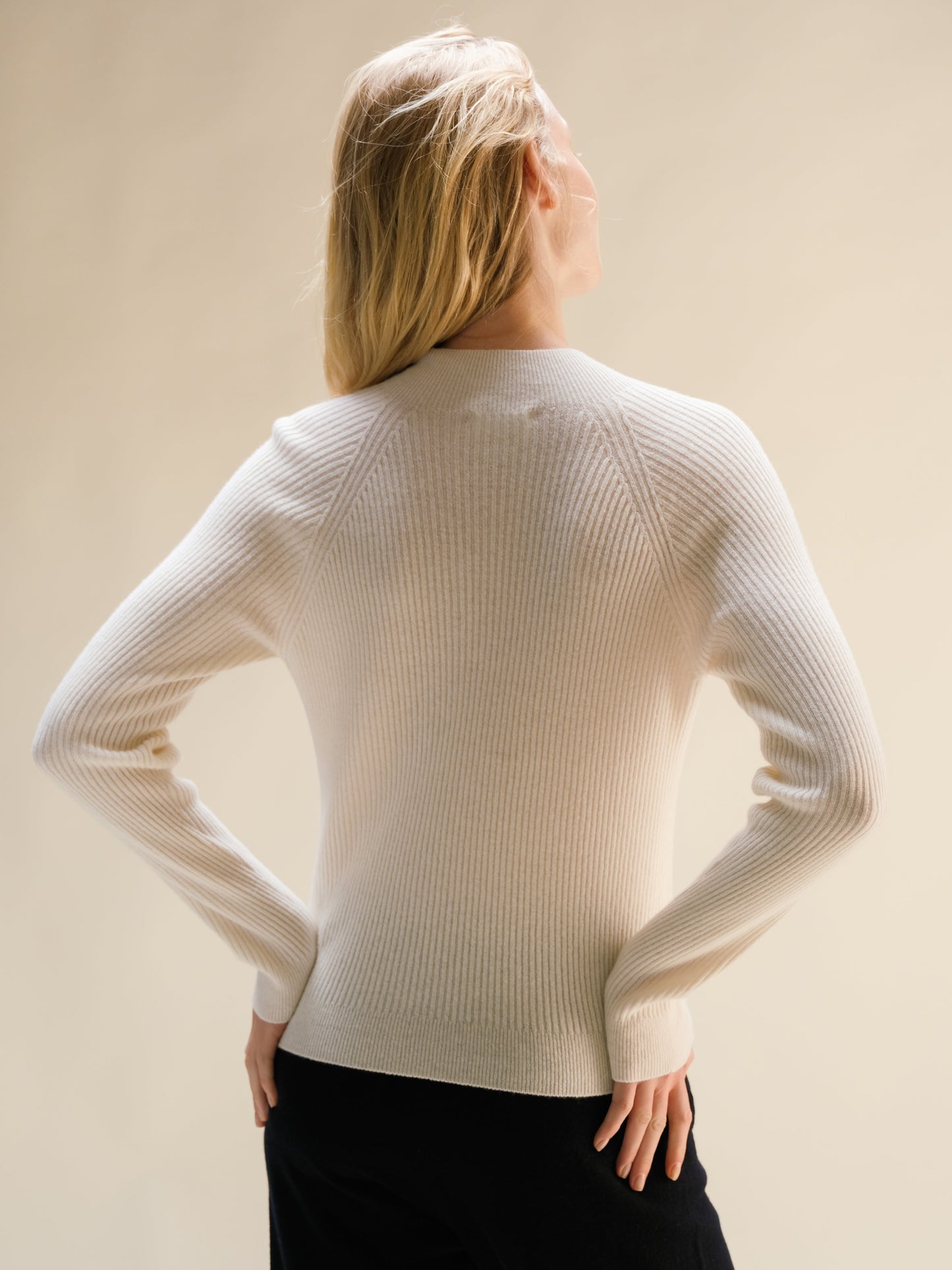 Model wearing cream ribbed cashmere mix mock neck sweater, back view with hands on hips.