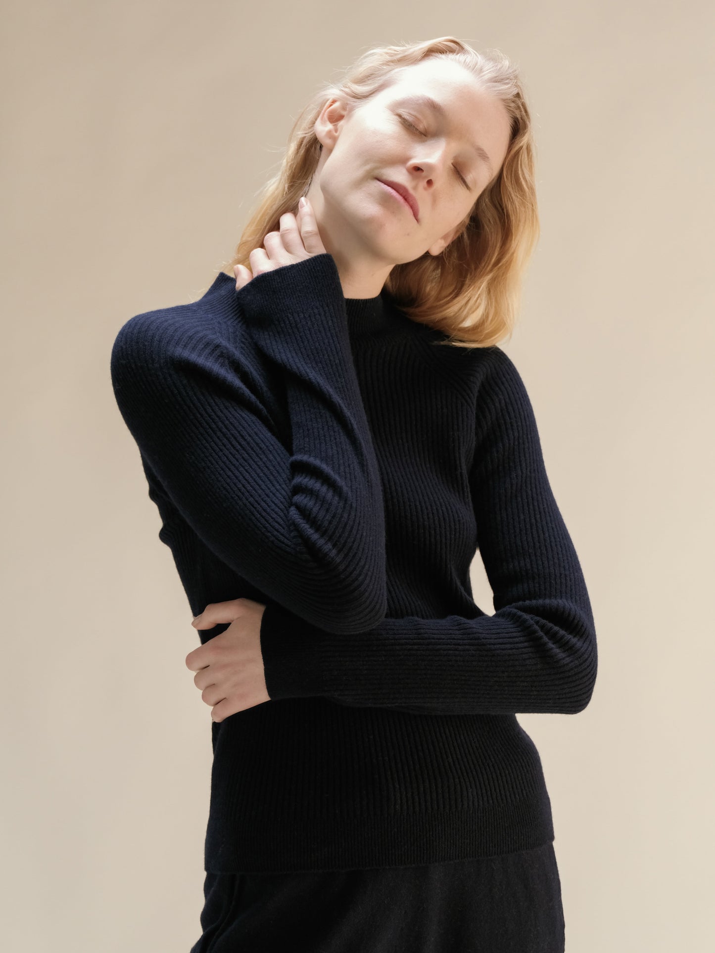 Model in navy cashmere mix ribbed mock neck sweater posing with eyes closed highlighting the soft knit.