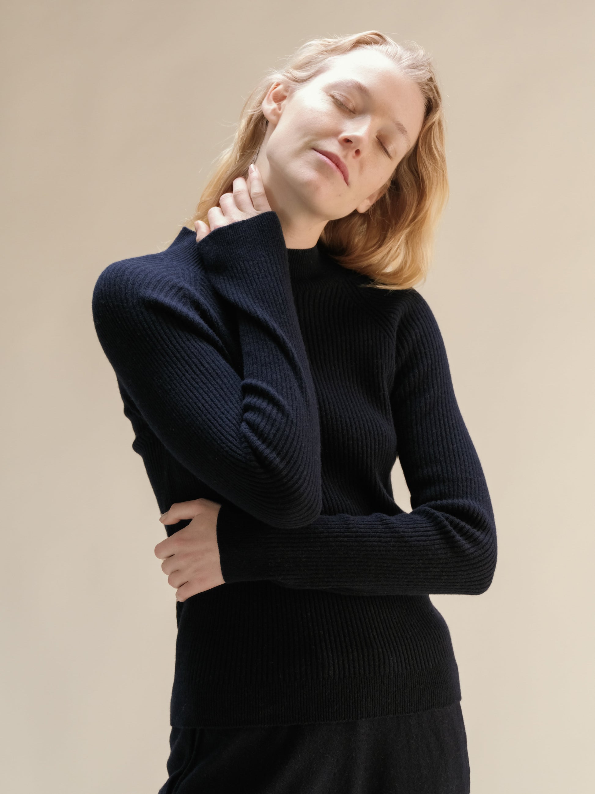 Model in navy cashmere mix ribbed mock neck sweater posing with eyes closed highlighting the soft knit.