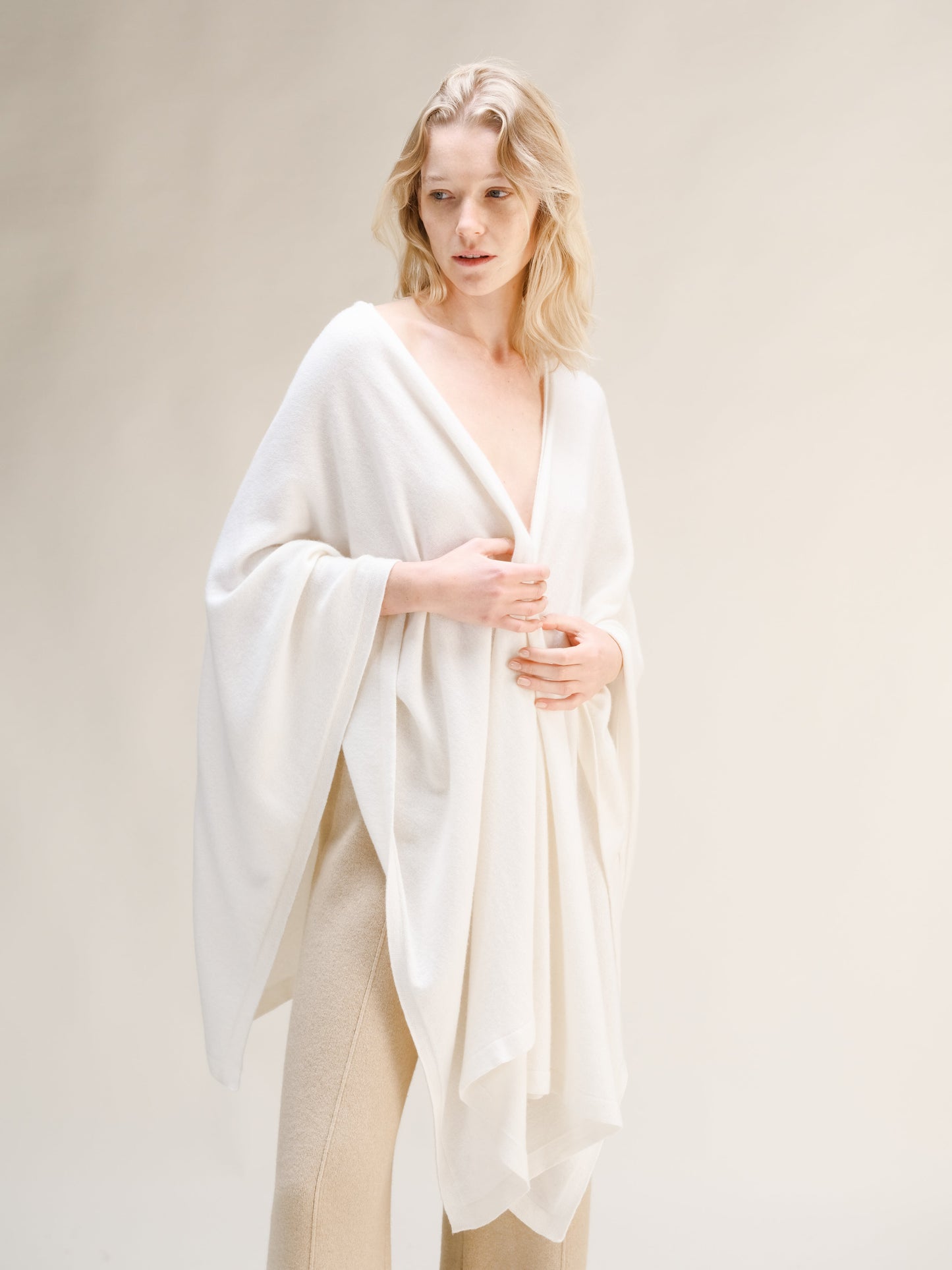 Model wearing a white cashmere mix maxi cape draped over shoulders with beige knit trousers.
