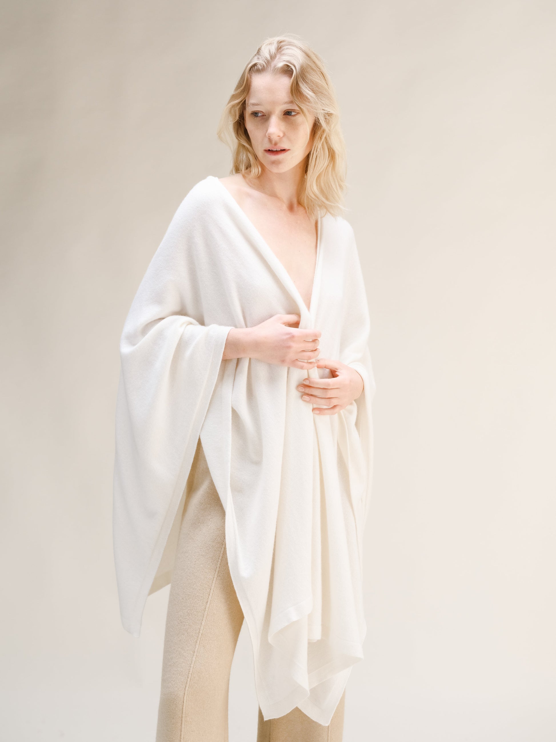 Model wearing a white cashmere mix maxi cape draped over shoulders with beige knit trousers.