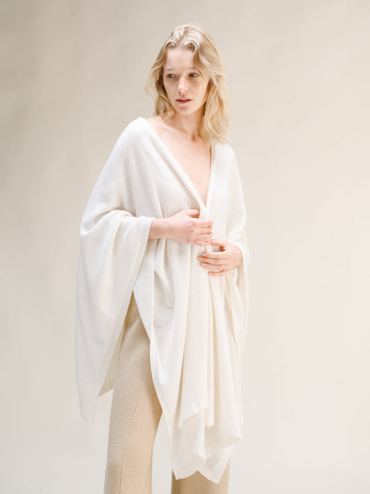 Model wearing a white cashmere mix maxi cape draped over shoulders with beige knit trousers.