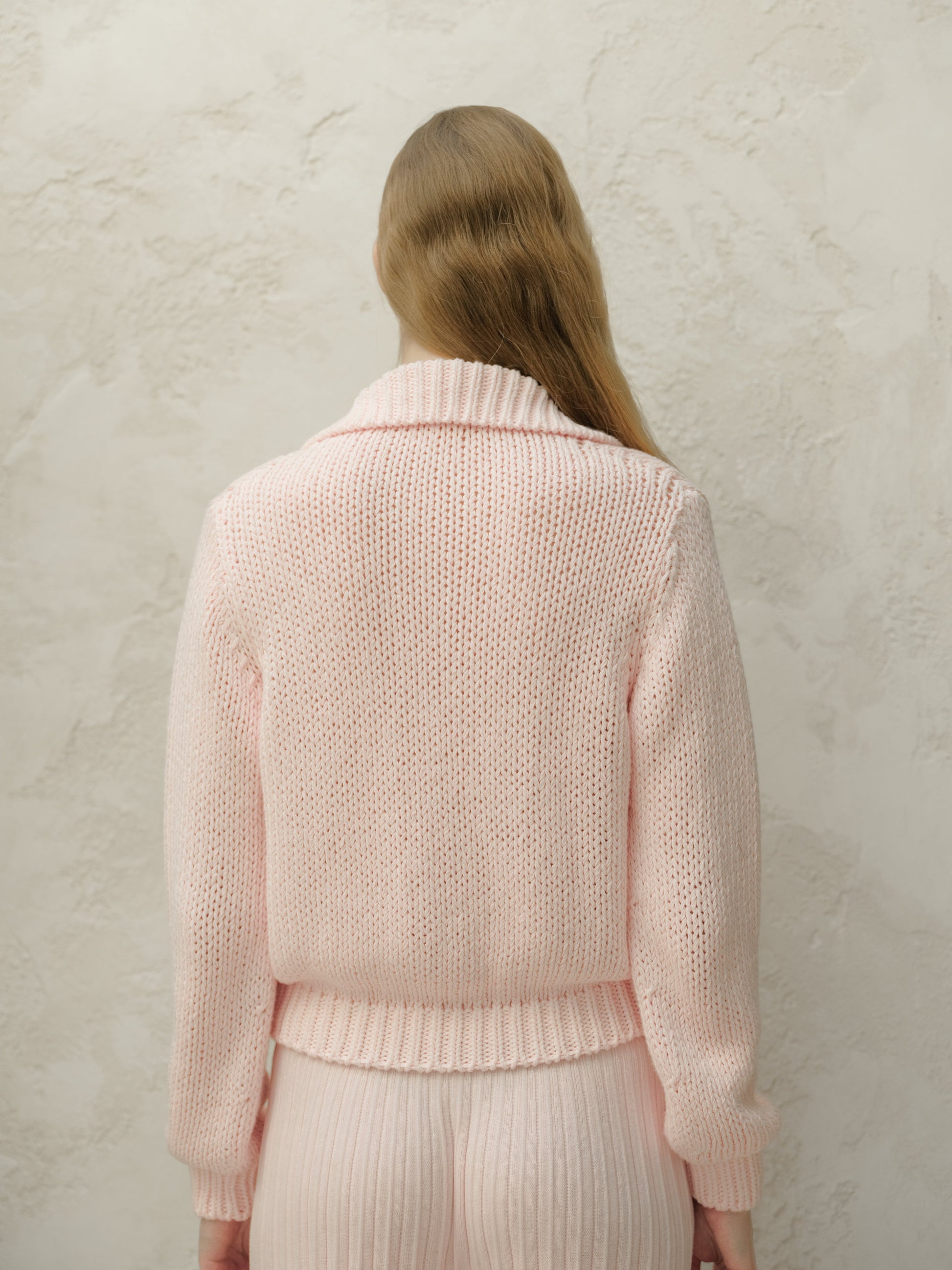 Back view of a model in a soft pink knit cardigan showing relaxed fit and ribbed hem against a textured wall