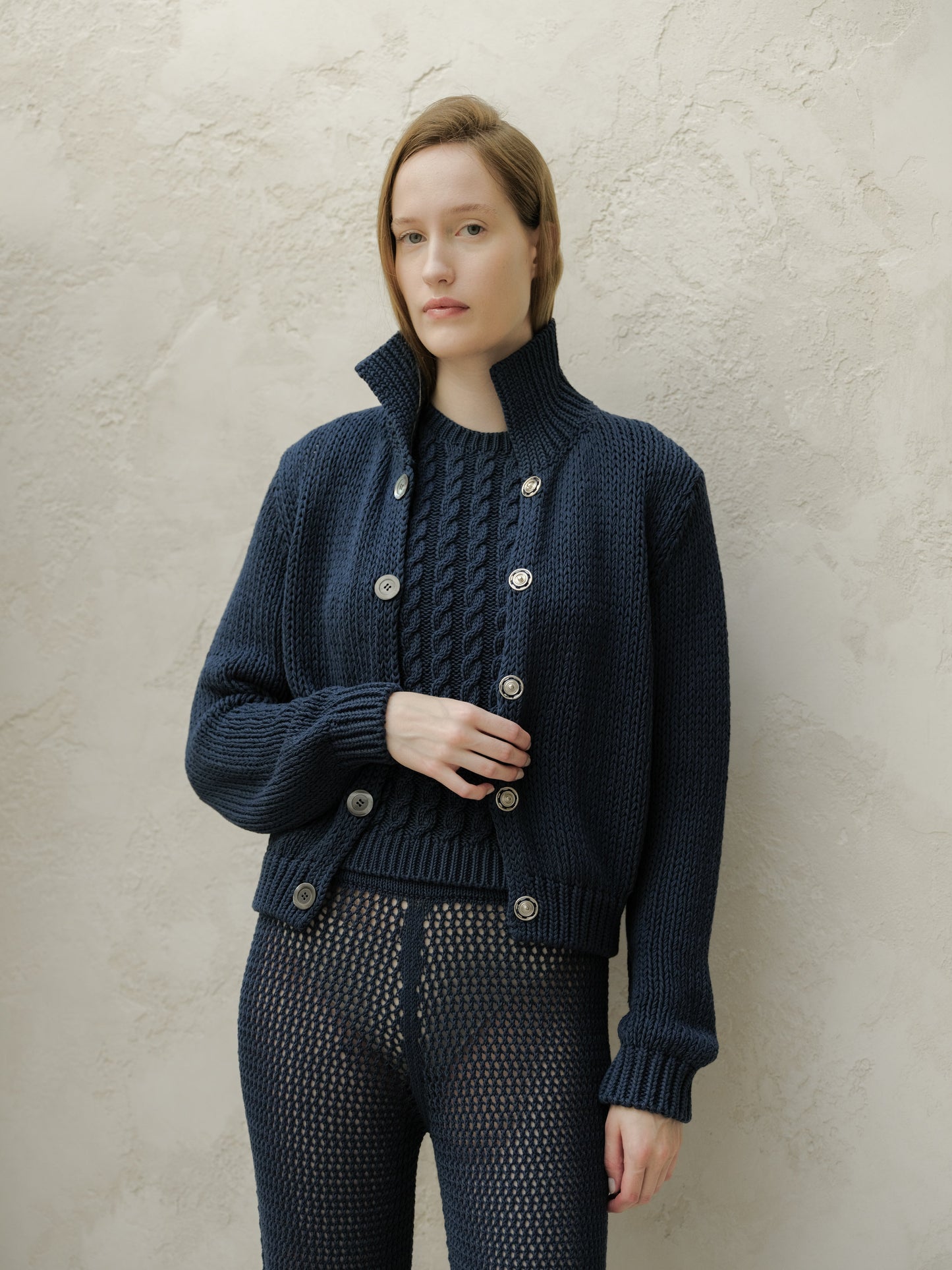 Woman wearing a navy chunky knit cardigan with a tall collar and large buttons stands against a textured beige wall.