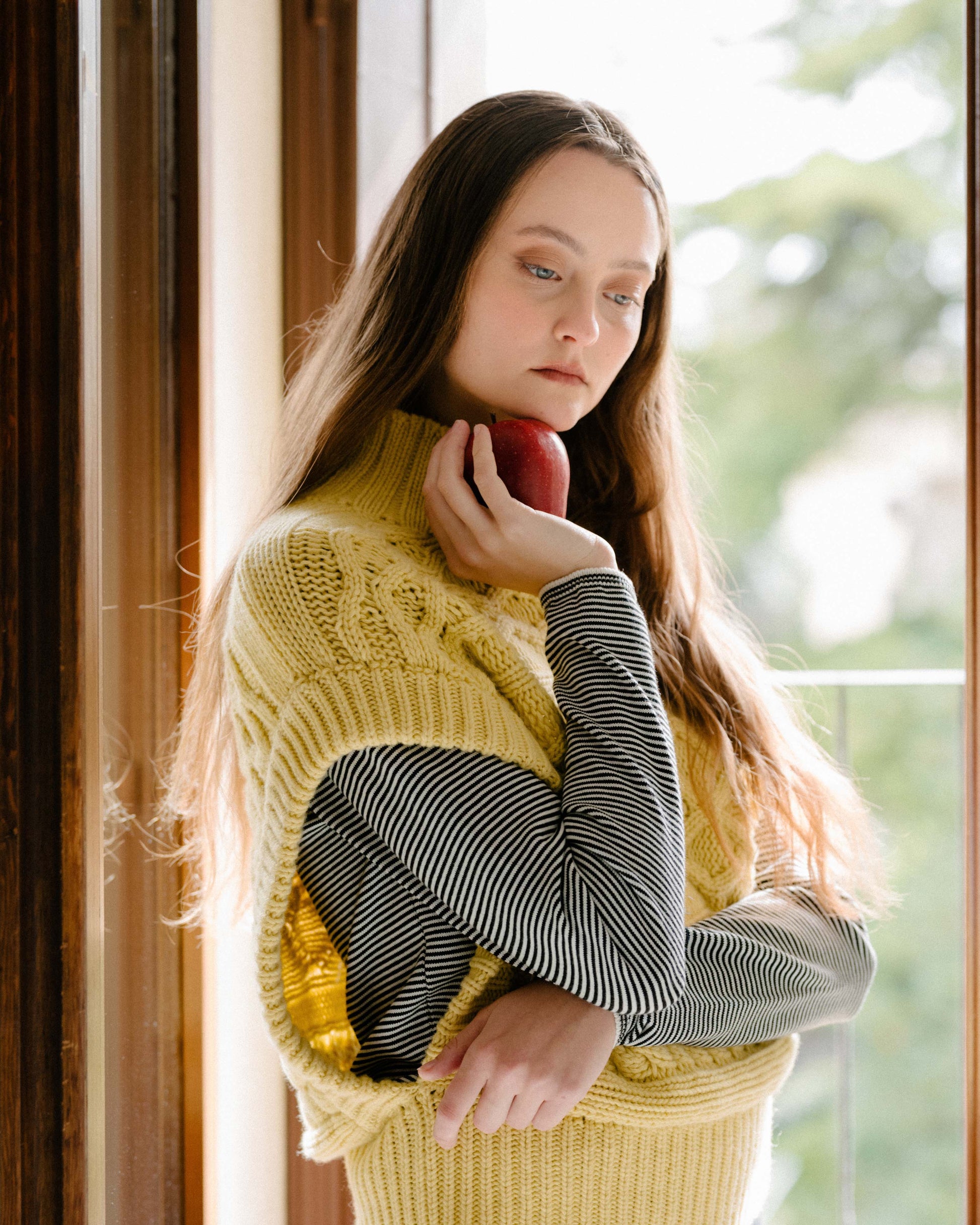 Female model wearing a cream cashmere mix maxi cable tunic over a striped top, holding a red apple near her face.