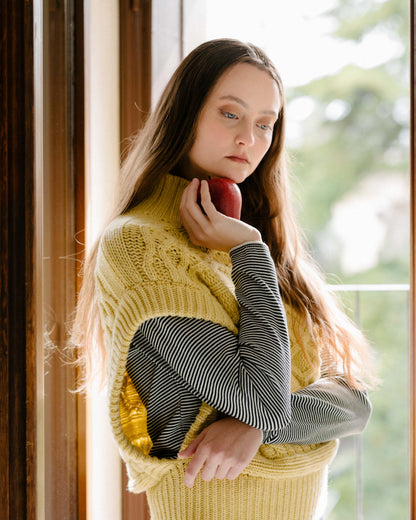 Female model wearing a cream cashmere mix maxi cable tunic over a striped top, holding a red apple near her face.