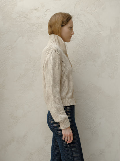 Side view of model in a beige knit bomber jacket with a high collar paired with navy openwork pants