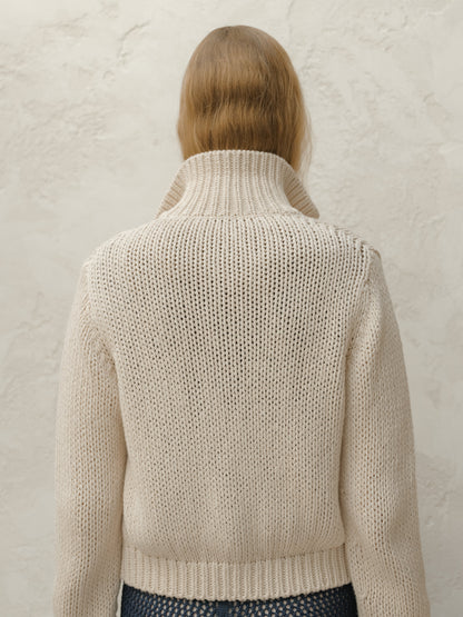 Back view of a model wearing a cream chunky knit bomber jacket with a ribbed hem against a textured light wall.